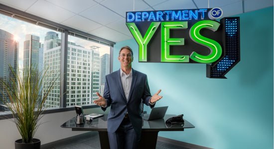 Man standing in front of desk in office with sign behind him that says Department of Yes